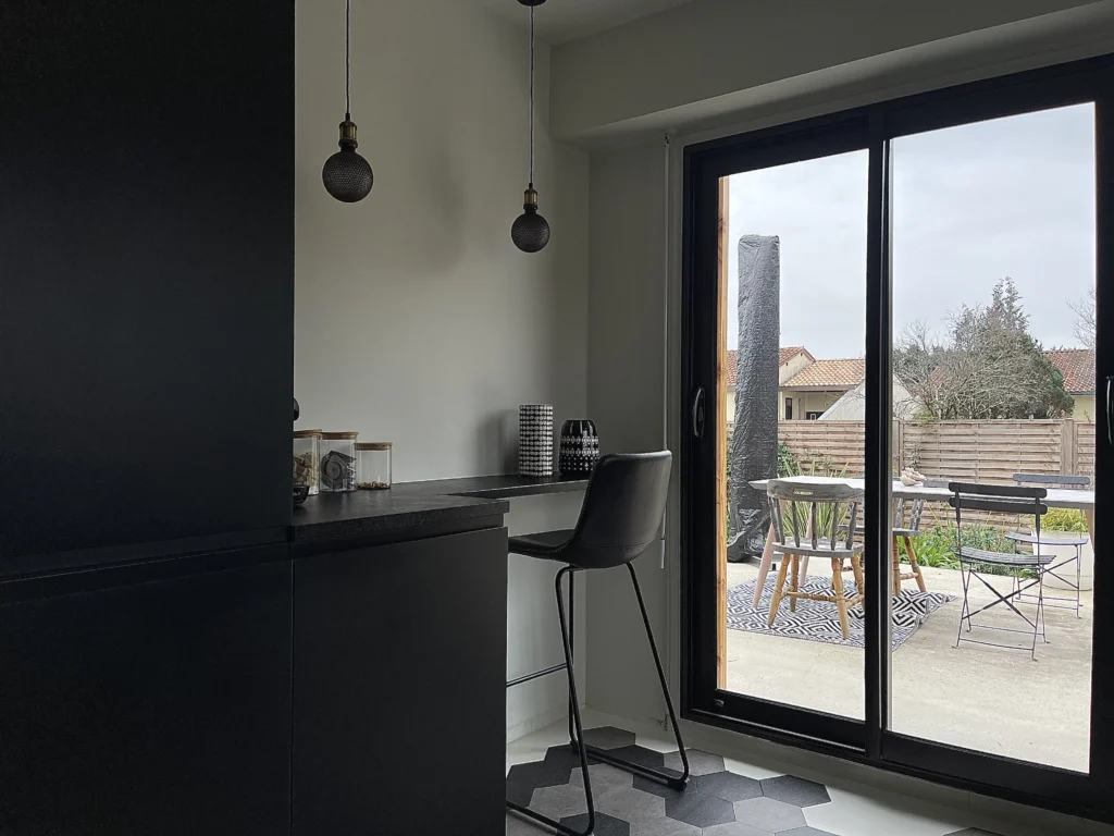 Coin repas dans une cuisine au style industriel avec tabouret haut noir, suspensions style atelier, et une porte-fenêtre donnant sur une terrasse extérieure